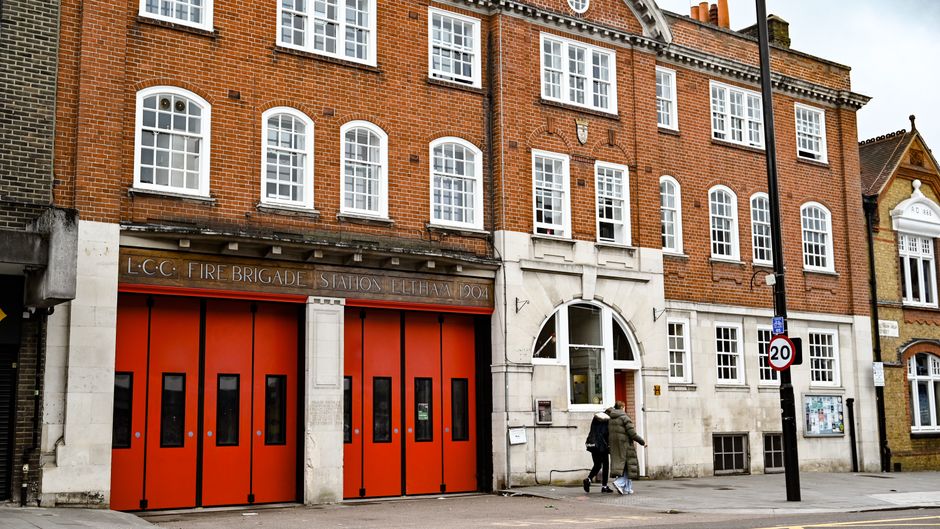 The exterior of a multi-storey brick building with white-framed windows and a prominent white stone entrance archway, situated in an urban area. In front of the building, there are two large orange firefighting doors labeled 'LCC Fire Brigade Station Eltham', indicating a fire station. The street scene includes a black lamppost with a 20 mph speed limit sign attached, as well as a notice board and other signage. Two individuals are walking past the entrance, one wearing a dark coat and carrying a bag, and the other in lighter clothing. The pavement is clean, and the lighting suggests daytime conditions. This setting is part of a property where house removals and home relocation services offered by Eltham Man and Van might be conducted, with the scene capturing the initial steps of loading or preparing furniture and packing materials for transport to a new address.