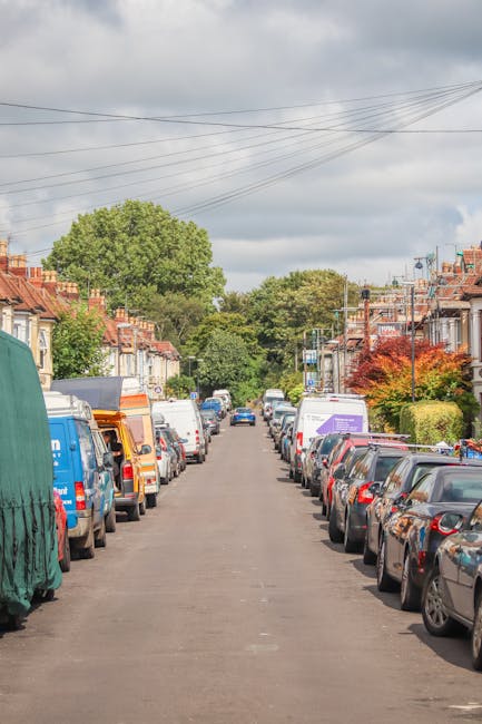 A narrow residential street with parked cars lining both sides, including compact hatchbacks, larger estate vehicles, and commercial vans. Several vehicles are covered with protective tarps or plastic wraps, indicating preparations for a home relocation or furniture transport. The street features typical terraced houses with brick facades and small front gardens, with some houses displaying potted plants or hedges. Utility poles and overhead power lines run along the street, while trees with lush green foliage and a cloudy sky are visible in the background. The scene suggests a quiet, densely built neighbourhood suitable for a professional removals service like Eltham Man and Van handling furniture moving and packing within limited space and narrow access properties. The environment is calm, showcasing the logistical challenges of loading and parking during house removals in this residential area, consistent with the process of home relocation as seen in the context of the Eltham High Street removals guide for narrow access properties.