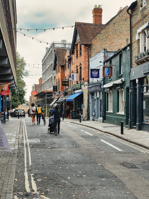 A narrow residential street with parked cars lining both sides, including compact hatchbacks, larger estate vehicles, and commercial vans. Several vehicles are covered with protective tarps or plastic wraps, indicating preparations for a home relocation or furniture transport. The street features typical terraced houses with brick facades and small front gardens, with some houses displaying potted plants or hedges. Utility poles and overhead power lines run along the street, while trees with lush green foliage and a cloudy sky are visible in the background. The scene suggests a quiet, densely built neighbourhood suitable for a professional removals service like Eltham Man and Van handling furniture moving and packing within limited space and narrow access properties. The environment is calm, showcasing the logistical challenges of loading and parking during house removals in this residential area, consistent with the process of home relocation as seen in the context of the Eltham High Street removals guide for narrow access properties.