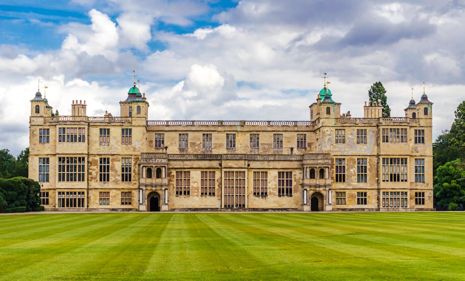 A large historic stone building with multiple rectangular windows and ornate architectural details, situated behind a well-maintained, expansive green lawn. The structure features four prominent corner towers topped with small green domes and gold weather vanes, while the central section displays symmetrical windows and a large arched doorway. The scene is set outdoors under a partly cloudy sky, with trees and shrubbery surrounding the property. In the context of house removals and home relocation, [COMPANY_NAME] may be involved in the process of carefully packing, transporting, and loading furniture and belongings from such a listed building, using appropriate equipment like trolleys and vans to ensure safe and efficient furniture transport during a house move near historic sites like Eltham Palace.