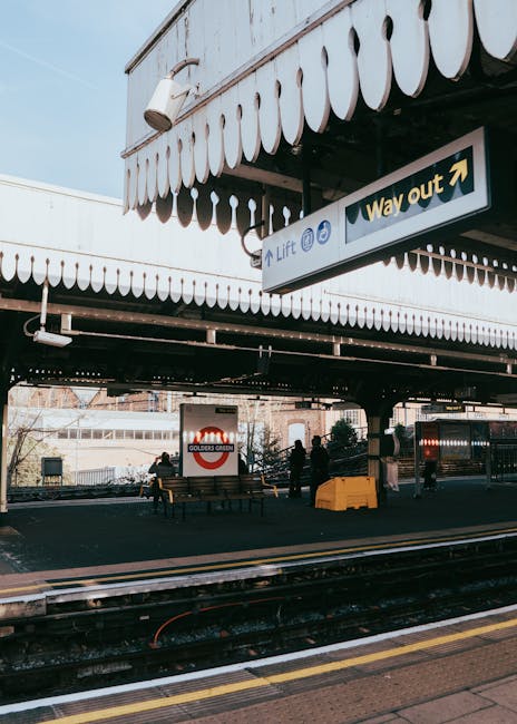 A view of the outdoor area at New Eltham station in London, showing a covered loading zone with a white canopy featuring decorative scalloped edging. Several black metal benches and a large white sign with the Golder's Green underground station logo are visible, along with a few silhouetted figures waiting or walking nearby. The area is paved with asphalt and located adjacent to railway tracks with yellow safety lines along the platform edge. A blue sign with white text indicates an exit labeled 'Way out,' and a smaller sign points to a lift. In the background, parts of the station building and surrounding urban environment can be seen. This scene illustrates a typical station loading and drop-off zone relevant to home relocation and furniture transport services offered by Eltham Man and Van, emphasizing the logistics involved in moving items to or from a station area.