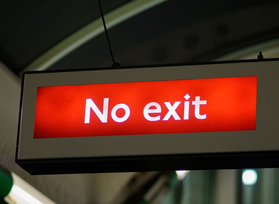 A rectangular illuminated sign hanging from the ceiling indoors displays the message 'No exit' in bold white letters on a red background. The sign is framed by a white border and suspended by two black cords. The background includes a blurred view of an indoor environment, possibly a station or loading area, with visible ceiling structures and lighting. The lighting in the scene is even, highlighting the sign clearly. This image illustrates a typical signage used in transportation hubs or loading zones, relevant to house removals and moving logistics, such as directing or restricting access at a loading or unloading point near New Eltham station, as indicated by the page title and context on elthammanandvan.com.