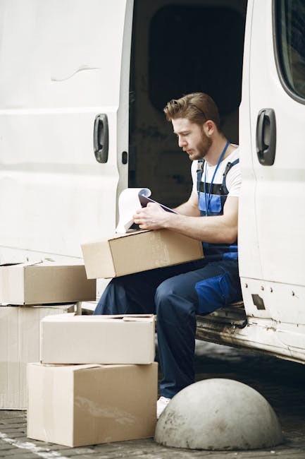 A woman sitting cross-legged on a wooden floor surrounded by multiple large cardboard moving boxes, some sealed with red tape and others open. She is wearing casual clothing, including a white tank to
